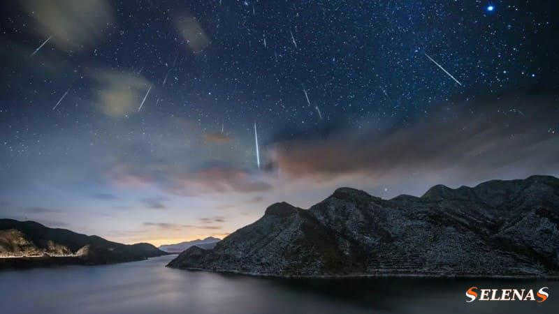 The Geminid meteor shower occurs over a large lake surrounded by hills. Shooting stars streak across the starry sky.