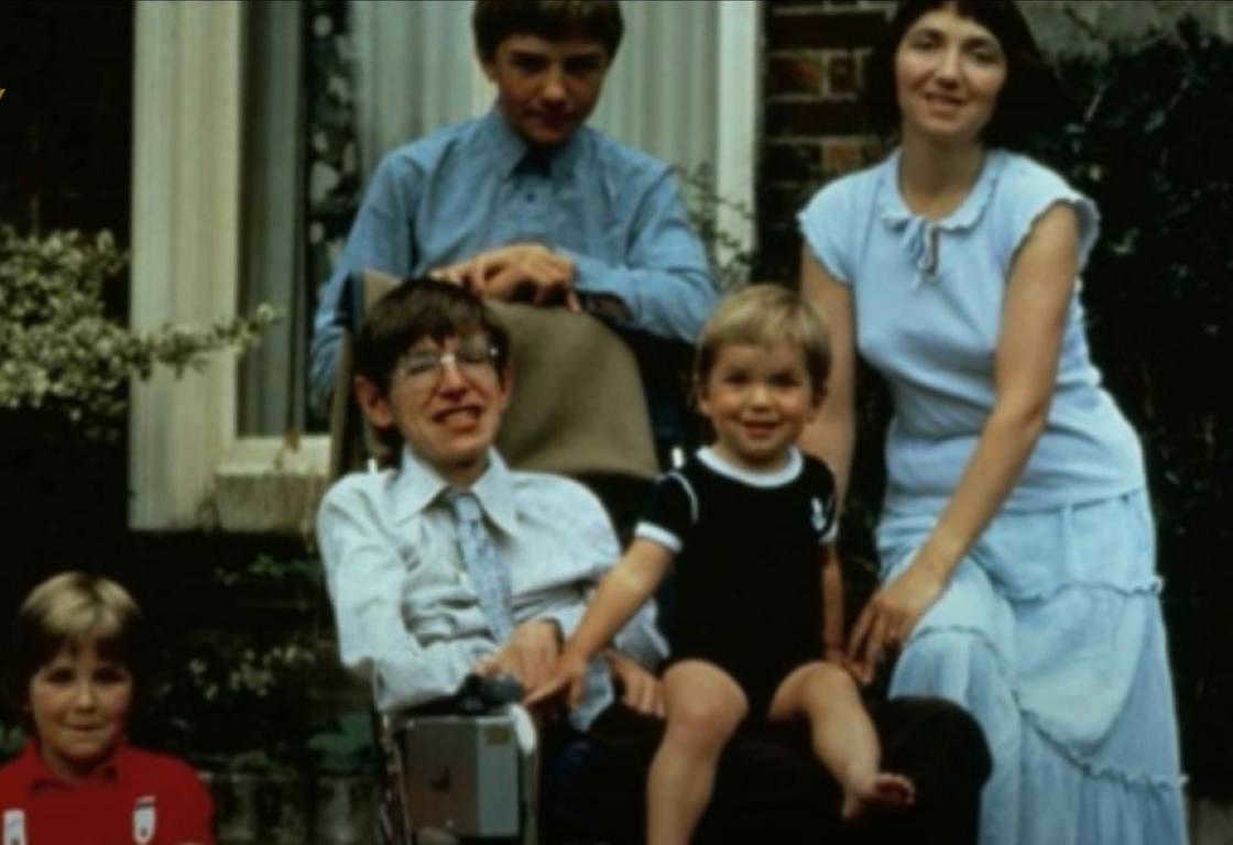 A man with glasses with a little boy on his lap, next to a woman, an adult boy and a girl