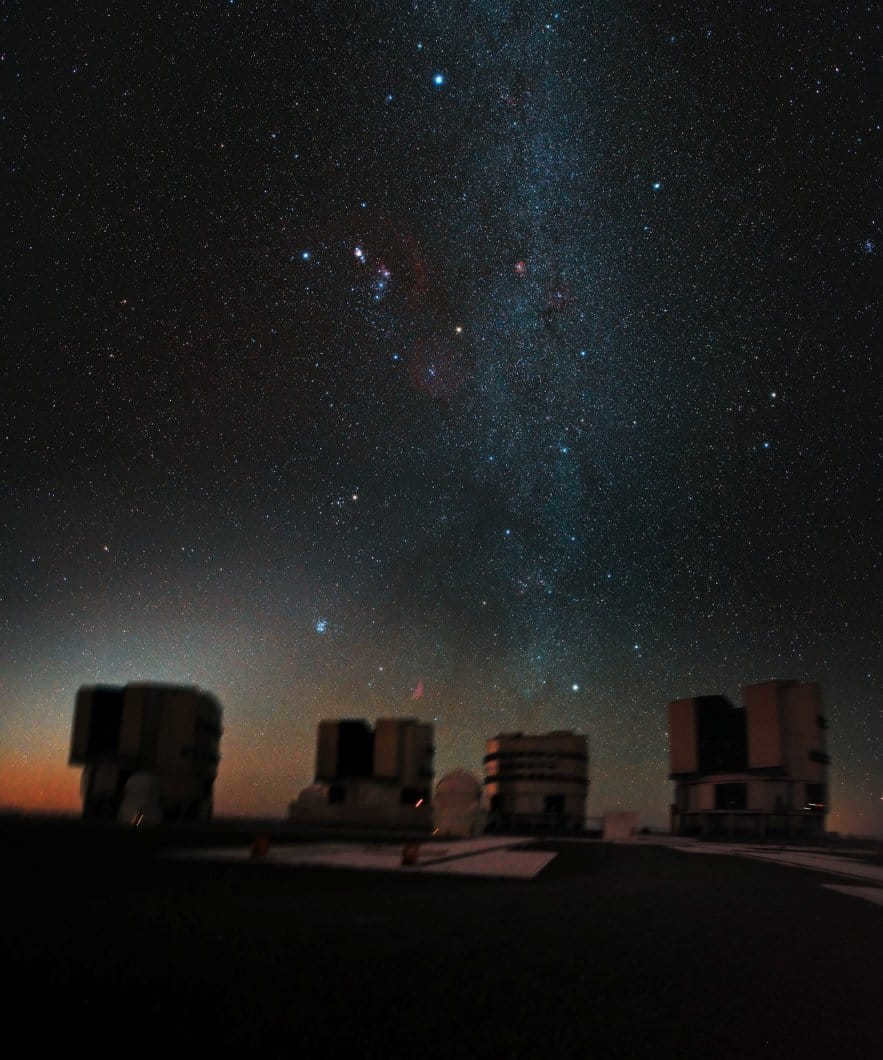 Orion, observed by the Very Large Telescope. The instrument is in the Southern Hemisphere, so Orion is head down here, as if trying to plunge into the Atacama Desert. At night, 4 giant 8.2 meter telescopes raise their lenses into the sky to study the universe. Above the telescope stretches the band of the Milky Way with the bright star Capella (just above the telescope). Above and to the left is Orion's belt and sword with the Orion Nebula between blue Rigel and orange Betelgeuse. In the middle is the red Rosetta Nebula and above is the bright star Sirius